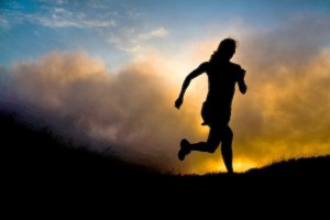 A woman runs on a trail silhouetted by a setting sun shrouded by fog.